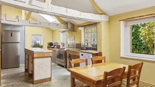 Salle à manger lumineuse en jaune vif avec table et chaises en bois, cuisine ouverte avec électroménagers en acier inoxydable, poutres apparentes
