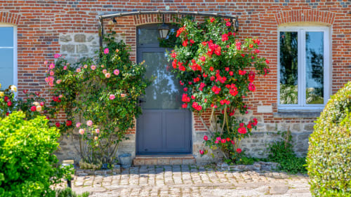 Des roses grimpantes rouges encadrent la porte bleu foncé du cottage à L'etoile De Forges, Longmesnil, avec façade en brique et jardins soignés.