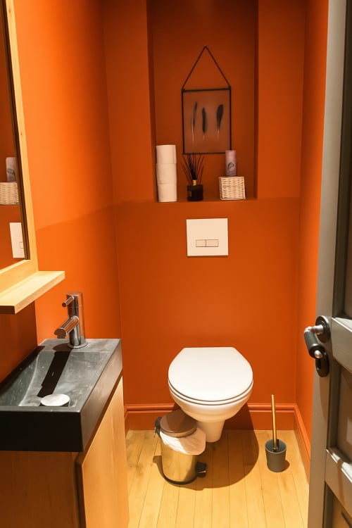 Salle de bain moderne aux murs orange vif, toilettes blanches, lavabo sombre et étagère murale décorée à l'hôtel Maunoury Citybreak