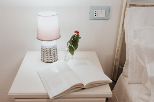 Tavolino da notte in appartamento italiano con lampada, libro aperto e fiore rosso in vaso