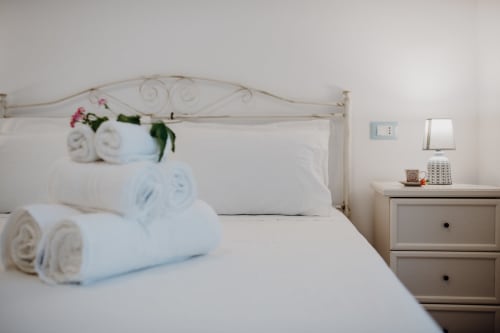 Camera da letto luminosa con letto bianco, asciugamani arrotolati, fiori rosa, comodino con lampada e decori all'appartamento Il Porticicolo Vacanze