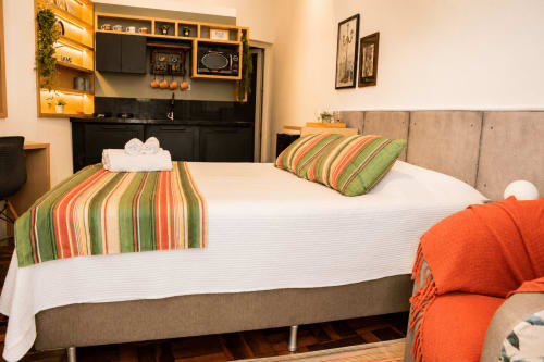 Quarto de loft em Maringá com cama queen, almofadas listradas, cabeceira bege, kitchenette e vista para o jardim