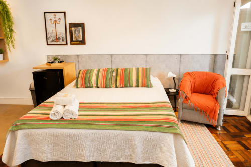 Quarto Veneto Loft no CSS em Maringá com roupa de cama listrada, cabeceira cinza e poltrona laranja