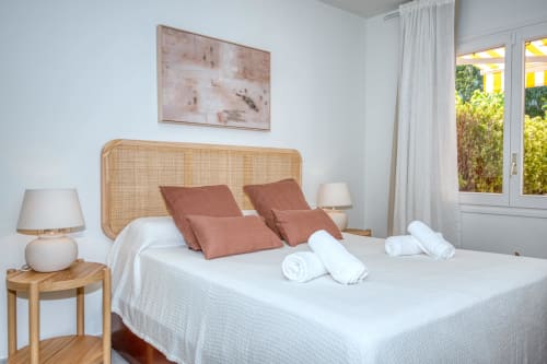 Modern bedroom with rattan headboard, white bedding, terracotta pillows, and garden view in Palafrugell apartment
