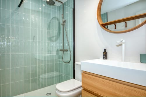 Modern bathroom in Palafrugell apartment with green tiled shower, white sink, wooden vanity, and round mirror