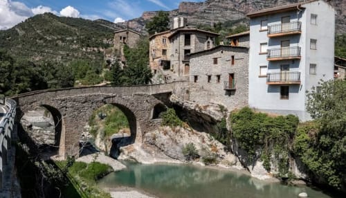 Puente medieval de piedra sobre río turquesa con vistas a montañas, edificios históricos y moderno edificio blanco de apartamentos