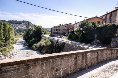 Puente de piedra con vista al río Montgat, edificios tradicionales de piedra y montañas Prepirenaicas al fondo