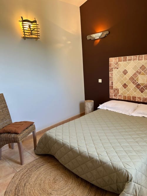 Chambre moderne à Vista D'oro à Monacia d'Aullène avec lit queen-size, tête de lit carrelée, fauteuil en rotin et éclairage chaleureux