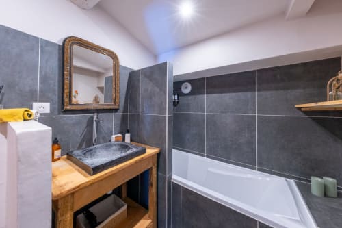 Salle de bain moderne avec carrelage sombre, meuble vasque en bois, miroir à cadre doré et baignoire au château de Grignan