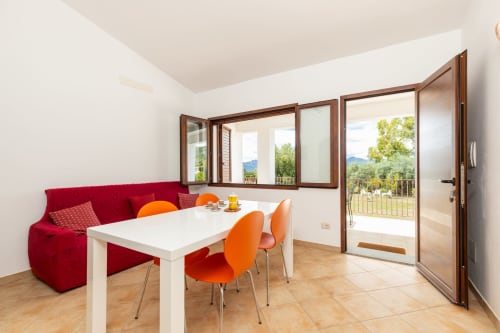 Modern apartment dining area with red sofa, white table, orange chairs, and glass doors overlooking garden at Agriturismo