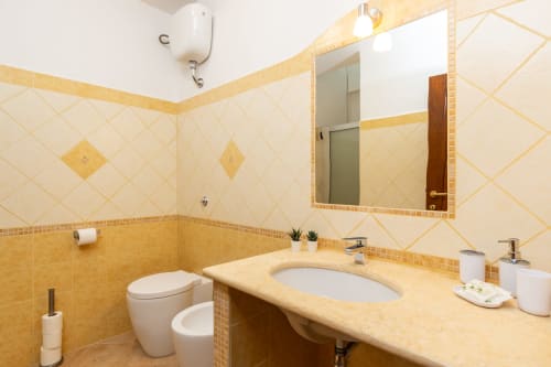 Bright bathroom with cream and gold tile work, vanity sink, toilet, mirror, and bidet in Agriturismo apartment, Bari Sardo