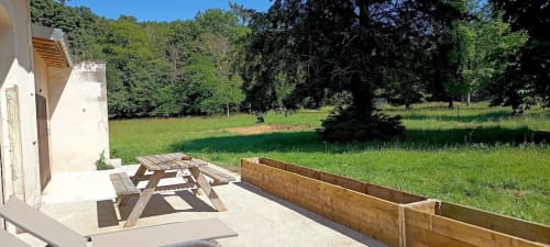 Table de pique-nique en bois sur terrasse avec bac de culture surélevé, bâtiment blanc de style cottage et verdoyants champs au Domaine Alga