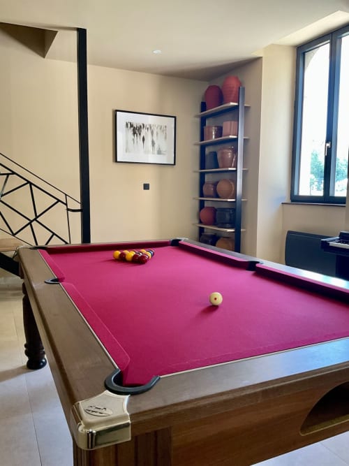 Salle de jeux lumineuse avec billard rose, boules prêtes au jeu, escalier en bois, étagères avec vases rouges et grandes fenêtres