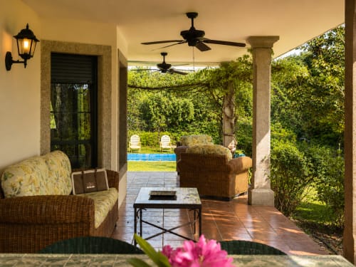 Porche cubierto con mobiliario de mimbre, ventiladores de techo y vistas a la piscina y jardín de la casa rural As Landras en Tomiño