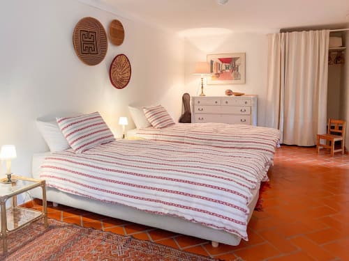 Quarto espaçoso na Quinta da Mata de Baixo com roupa de cama listrada em vermelho e branco, cômoda bege, cestos de palha na parede, e