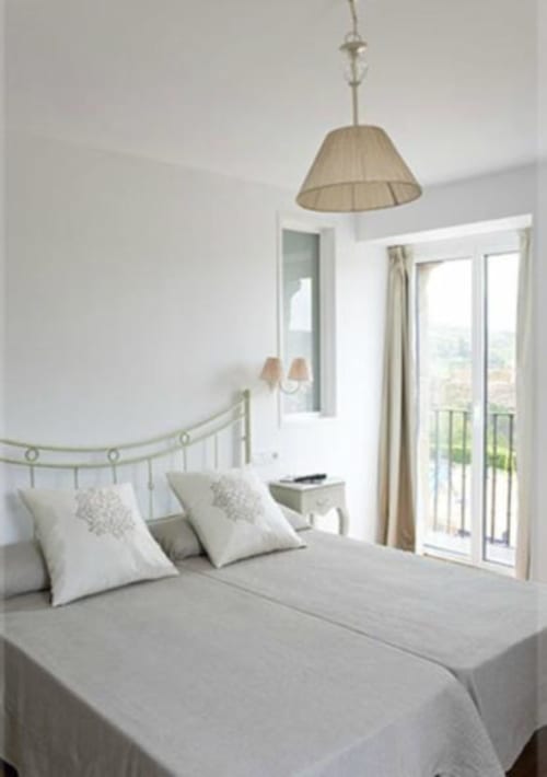 Dormitorio luminoso en Hostal Dona Blanca con lámpara colgante beige, cabecero metálico, almohadas blancas y puertas francesas que abren a