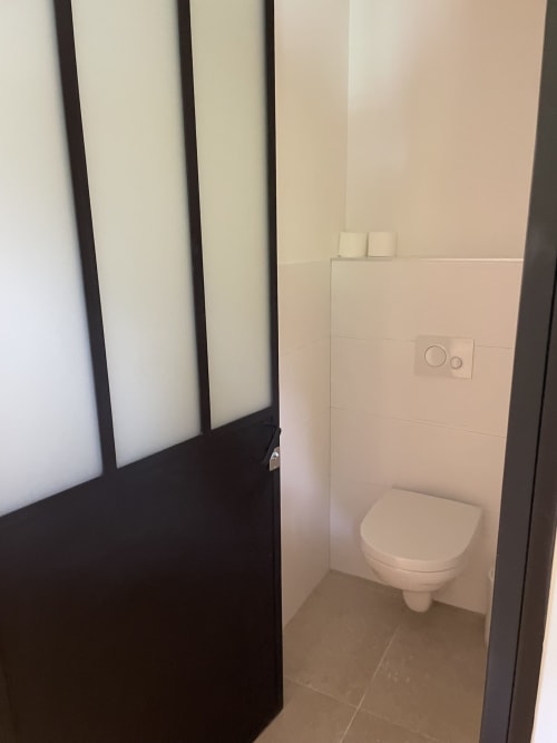 Toilettes modernes murales dans la salle de bain du gîte Les Pratges avec carrelage crème et encadrement de porte foncé à Figeac