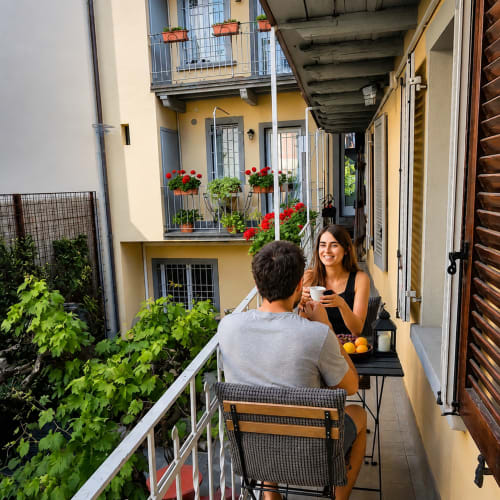 Coppia che gode del caffè su un soleggiato balcone degli Apartments Borgovivobg a Bergamo, circondata da fiori rossi e verde