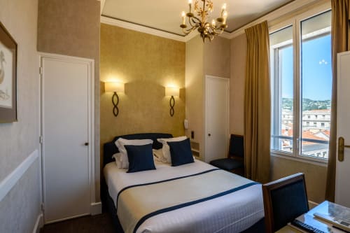 Elegante camera dell'Hotel Splendid Cannes con parete accent dorata, biancheria blu navy e vista sui tetti di Cannes