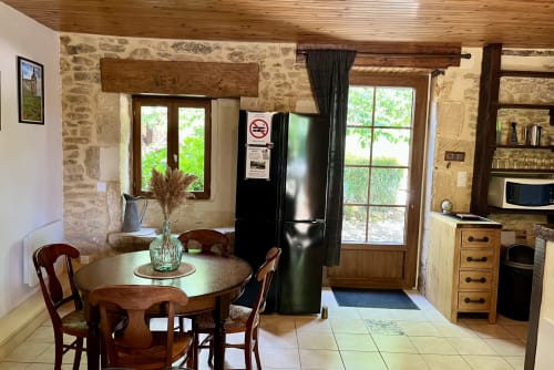 Coin cuisine-salle à manger rustique au Gîte La Vieille Ferme avec murs en pierre, table en bois, réfrigérateur noir et vue sur le jardin