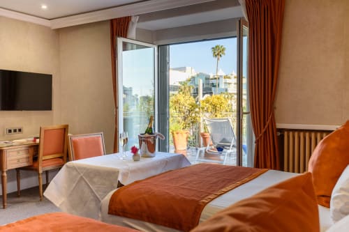 Camera lussuosa a Cannes con letti gemelli, terrazza con vista su palme e città, arredamento arancione e crema