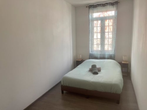 Chambre lumineuse aux Business Apparts Caudry avec lit blanc, tables de nuit en bois et vue sur la ville par fenêtre aux voilages