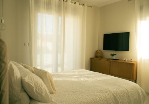 Chambre lumineuse à La Maison du Potier à Valras-Plage avec literie crème, TV murale, commode en bois et rideaux voilages
