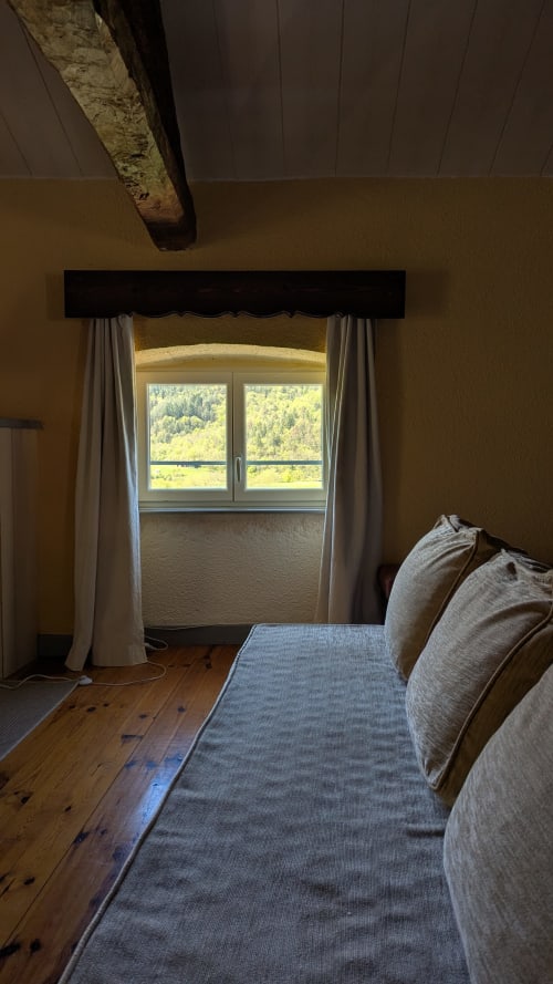 Camera da letto luminosa a Casa Orma con letto, finestra con vista sul giardino boschivo, tende color crema, pavimento in legno e travi a vista