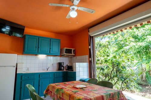 Cuisine lumineuse aux armoires turquoise, murs orange, ventilateur de plafond, table à manger avec nappe colorée, vue sur le jardin aux Gîtes