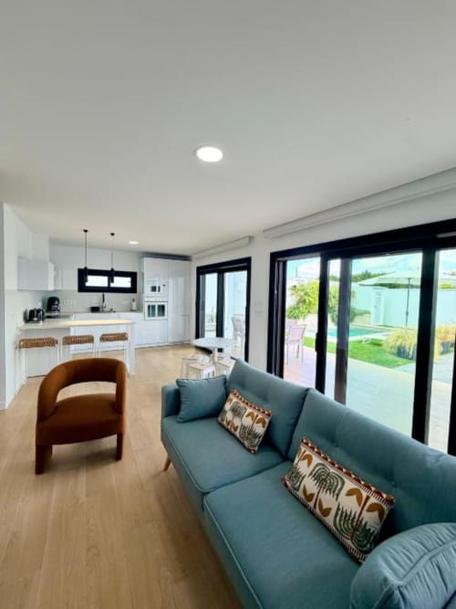 Sala de estar en villa moderna de planta abierta con sofá turquesa, sillón marrón, cocina blanca y puertas de vidrio con marco negro que se abren a