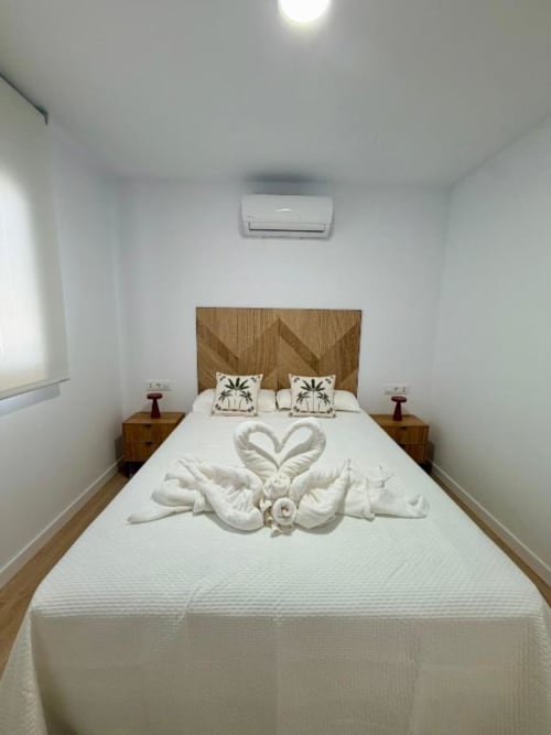 Elegante dormitorio de villa en Barrosa Home con sábanas blancas, cisnes decorativos de toallas, cabecera de madera y aire acondicionado