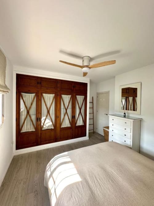 Dormitorio moderno en Villa Faro, Barrosa Home Hozanejos, con armario de madera, cómoda blanca, ventilador de techo y vistas al jardín