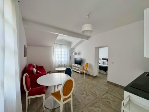 Salon lumineux avec canapé rouge, table blanche ronde, chaises jaunes, téléviseur et accès à la chambre au Domaine de Bize