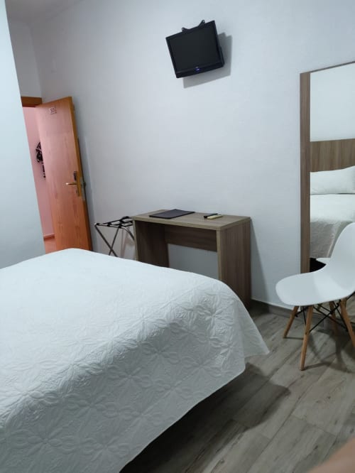 Dormitorio doble de Hotel La Posada con cama blanca, escritorio de madera, silla, televisor de pantalla plana y puerta rosa en España