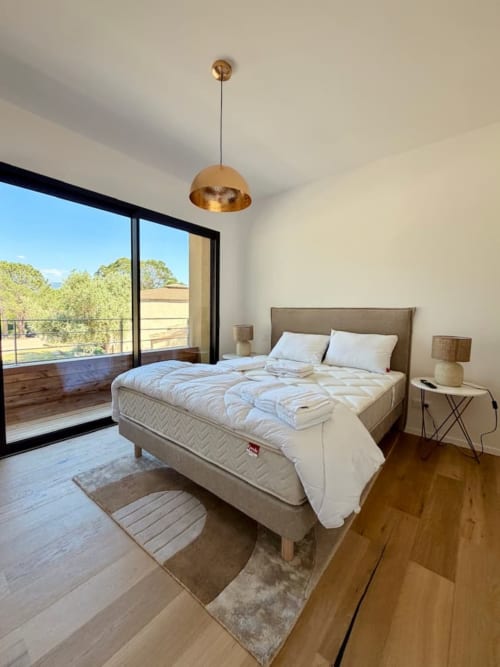 Chambre moderne aux Villas d'A Storia à Porto-Vecchio avec lit, luminaire doré et balcon donnant sur la verdure
