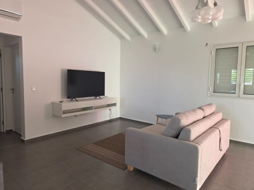 Salon de villa moderne à Maly Blue Location avec téléviseur écran plat, canapé gris, poutres apparentes et portes jalousies à Le Moule