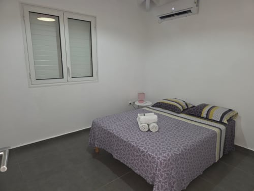 Chambre spacieuse avec lit double, fenêtre à volets blancs, climatisation et serviettes roulées à la villa Maly Blue Location au Moule