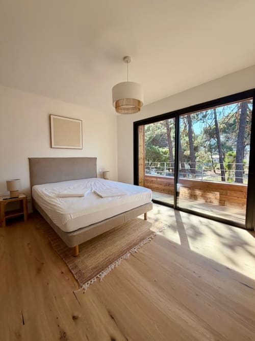 Chambre lumineuse avec lit queen, tête de lit grise, parquet et portes-fenêtres ouvrant sur la forêt aux Villas