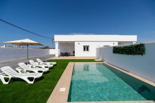 Villa moderna con piscina turquesa, tumbonas blancas, jardín verde y patio cubierto en Barrosa Home, España