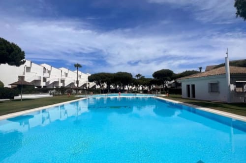 Piscina de aguas cristalinas en el resort Barrosa Home con edificios blancos y jardines diseñados bajo cielo azul