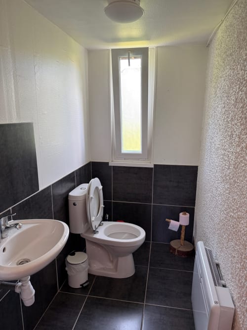 Salle de bains moderne du Chalet Cossu avec carrelage noir, lavabo blanc, toilettes, fenêtre et radiateur de chauffage