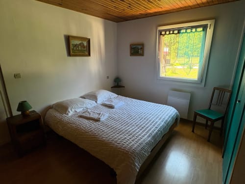 Bright bedroom in Gîte Jardin cottage with double bed, garden view window, wooden ceiling, and turquoise door