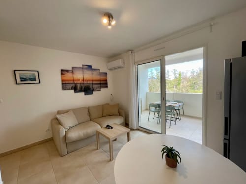 Modern apartment living room at Résidence Marina di Bravone with beige sofa, balcony overlooking forest, white dining table