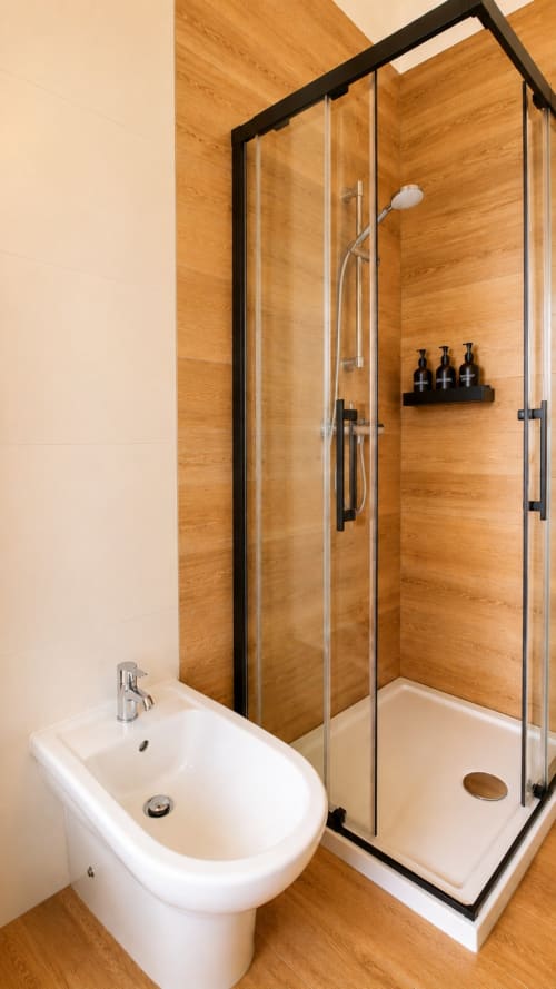 Bagno moderno con bidet e wc bianchi, doccia con box in vetro e rivestimento in legno, rubinetteria nera e accessori