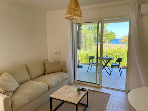 Monolocale moderno a Marina di Bravone con divano beige, tavolo in legno, lampada a sospensione intrecciata e accesso alla terrazza con vista mare