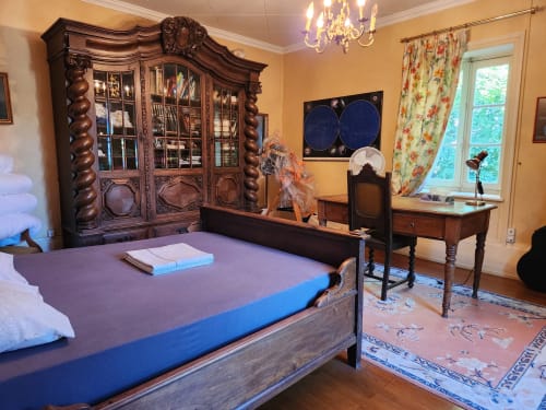 Chambre élégante dans villa française avec mobilier en bois ouvragé, lit bleu, cheminée et vue sur le jardin à travers des rideaux fleuris