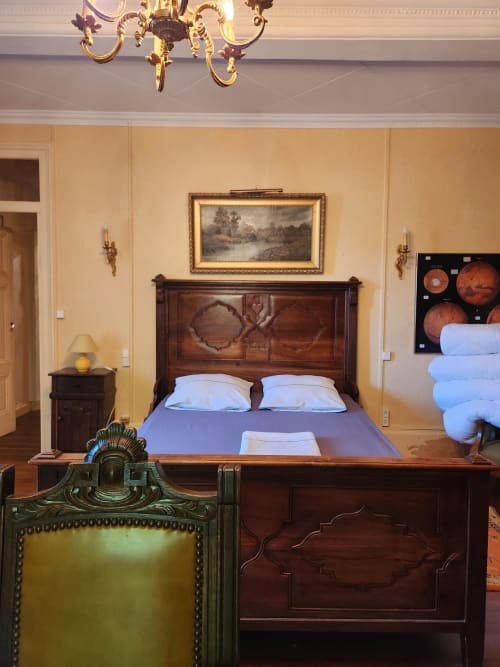 Chambre élégante à la Jolie Maison à Charly avec lit en bois travaillé, linge bleu, lustre doré et murs jaune pâle
