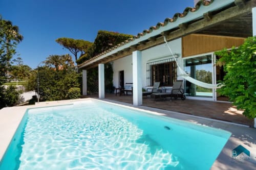 Área de piscina Barrosa con agua turquesa, villa blanca moderna, terraza de madera, tumbonas y jardín mediterráneo