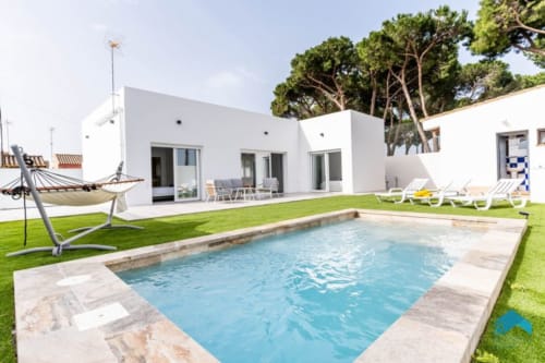 Villa moderna blanca de Barrosa Home con piscina rectangular, tumbonas, hamaca, césped exuberante y pinos en España
