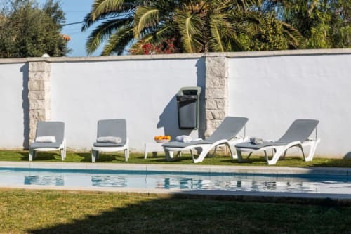 Zona de descanso junto a la piscina de Barrosa Home con tumbonas grises, pared blanca, palmeras y agua cristalina en España